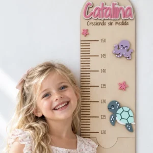 Niña sonriente junto a su medidor de altura infantil de madera personalizado con el nombre Catalina en rosa y la frase «Creciendo sin medida», con el pulpo, la tortuga y las estrellas de mar